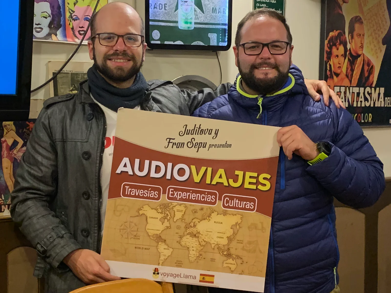 Juditova and Fran Sepu holding an audioViajes poster featuring a world map